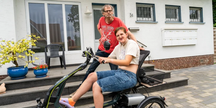 Ein helles Elektrodreirad mit gr&uuml;nem Schriftzug "Van Raam" steht auf einem gepflasterten Bereich vor einem wei&szlig;en Wohnhaus. Eine Person sitzt auf dem Sitz, tr&auml;gt Jeansshorts und ein helles T-Shirt, die F&uuml;&szlig;e ruhen auf den Pedalen. Eine zweite Person in rotem Shirt steht daneben, vor einer Treppe mit blauen Blument&ouml;pfen und zwei schwarzen St&uuml;hlen. Im Hintergrund sind Fenster und Briefk&auml;sten sichtbar.