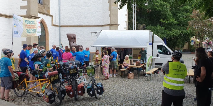 Versammlung von Menschen auf einem gepflasterten Platz vor einer wei&szlig;en Kirchenwand. Im Vordergrund stehen beladene Fahrr&auml;der, darunter ein gelbes Tandem. Personen tragen blaue und gelbe Westen und unterhalten sich. Rechts ein wei&szlig;er Pavillon und ein Transporter, dahinter mehrere Tische mit Materialien. Links h&auml;ngt ein Werbebanner. Im Hintergrund B&auml;ume und Geb&auml;ude.