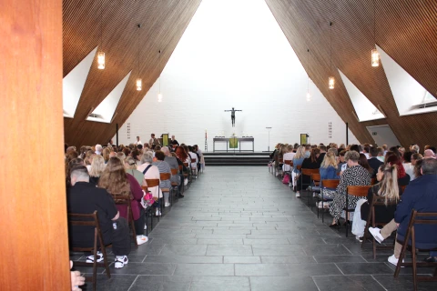 Innenansicht einer modernen Kirche mit holzverkleidetem, spitz zulaufendem Dach und h&auml;ngenden Lampen. Menschen sitzen auf Holzst&uuml;hlen in Reihen. Im Altarraum ein schwarzes Kruzifix vor einer wei&szlig;en Wand, ein Altar und Kerzenst&auml;nder.