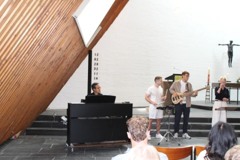 Gruppe von drei Musikern und einer S&auml;ngerin in einer modern gestalteten Kirche mit wei&szlig;er Ziegelwand und schr&auml;gem Holzdach. Links ein Klavier mit Noten, mittig ein Bassist und ein weiterer Musiker stehend, rechts die S&auml;ngerin am Mikrofon. Im Hintergrund ein schlichtes Holzkreuz, Stufen und eine brennende Kerze auf dem Altar.