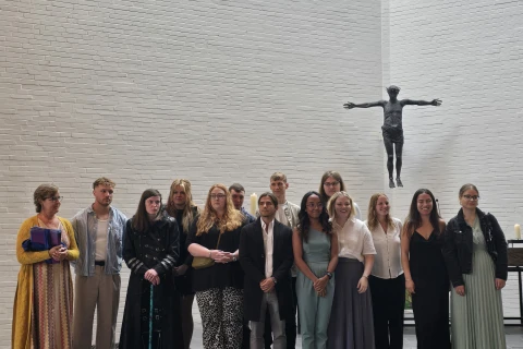 Gruppe von elf stehenden Personen auf grauen Stufen vor einer wei&szlig;en Backsteinwand. Eine schwarze Christusstatue mit ausgestreckten Armen schwebt an der Wand. Tageslicht str&ouml;mt durch diagonal verlaufende Fenster. Links und rechts auf dem Podest stehen einzelne Kerzen.