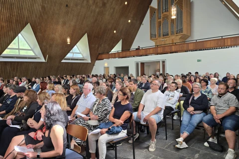 Eine gro&szlig;e Versammlung von Menschen, die auf St&uuml;hlen in einer Kirche mit moderner Holzarchitektur, schr&auml;gen Decken und gro&szlig;en dreieckigen Fenstern sitzen. Im Hintergrund ist eine Pfeifenorgel und eine Empore sichtbar. Alle Personen schauen nach vorne.
