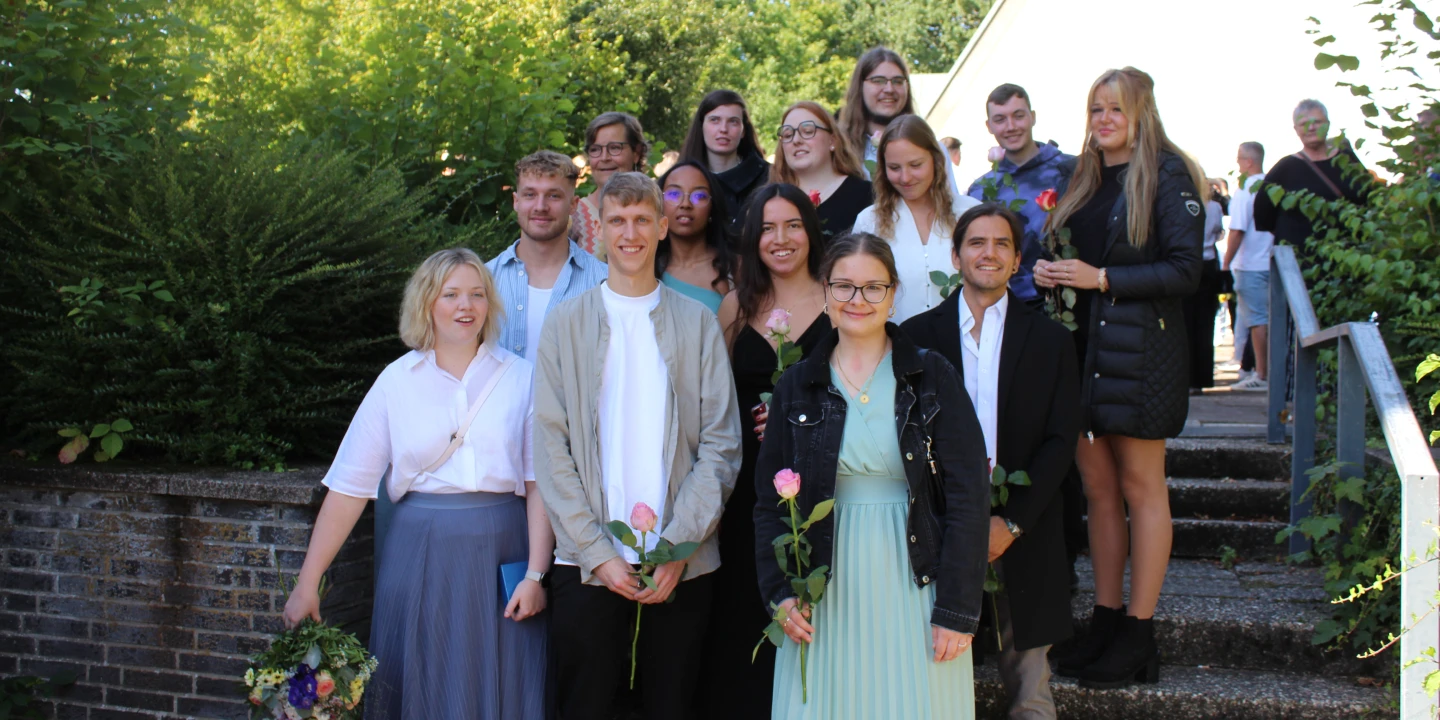 Personengruppe auf einer Treppe im Freien, umgeben von gr&uuml;nen B&auml;umen und einer niedrigen Ziegelmauer. Einige Personen halten Blumen in der Hand, eine Frau rechts tr&auml;gt eine schwarze Jacke und steht neben einem Gel&auml;nder. Im Hintergrund weitere Personen und Geb&auml;udeteile sichtbar.