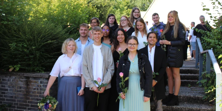 Personengruppe auf einer Treppe im Freien, umgeben von gr&uuml;nen B&auml;umen und einer niedrigen Ziegelmauer. Einige Personen halten Blumen in der Hand, eine Frau rechts tr&auml;gt eine schwarze Jacke und steht neben einem Gel&auml;nder. Im Hintergrund weitere Personen und Geb&auml;udeteile sichtbar.