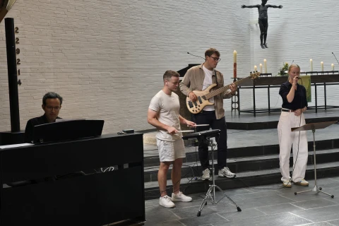 Vier Musiker in einer Kirche: links ein Klavierspieler an einem schwarzen E-Piano, daneben ein Mann mit Trommelsticks vor einem kleinen Schlagzeug, ein Mann mit E-Bass, und eine Frau mit Mikrofon auf einer dunklen Steinfl&auml;che, im Hintergrund Altar mit Kerzen und eine Christusfigur an der wei&szlig;en Ziegelwand.