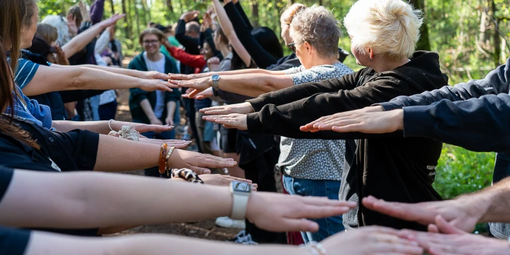 Menschen stehen in mehreren Reihen in einem Wald, strecken die Arme waagerecht nach vorne und ber&uuml;hren dabei fast die H&auml;nde der gegen&uuml;berstehenden Personen. Umgeben von gr&uuml;nen B&auml;umen und Sonnenschein.