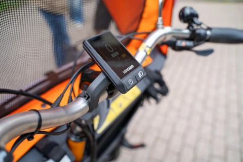 Lenker eines Lastenfahrrads mit integriertem Display. Das Display zeigt null Kilometer pro Stunde und ein Batteriesymbol. Der Lenker ist aus silbernem Metall, mit schwarzem Kunststoffgriff. Im Hintergrund sind orangefarbene Stoffseiten und ein Schutznetz erkennbar. Blasser, gepflasterter Untergrund.