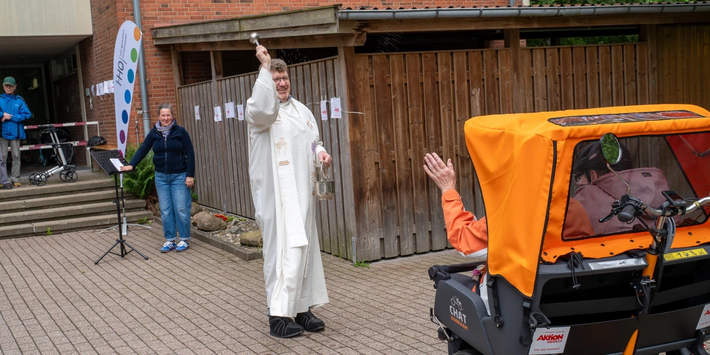 Ein Mann in cremefarbigem liturgischem Gewand steht auf einem gepflasterten Platz und hebt mit einer Hand ein Aspergill, w&auml;hrend er mit der anderen ein Metallgef&auml;&szlig; h&auml;lt. Rechts im Vordergrund ein orangener Fahrrad-Rikscha-Sitz mit einer Person darin, die einen Arm hebt. Im Hintergrund links ein Banner, eine Treppe und zwei weitere Personen. Holzwand und Backsteingeb&auml;ude im Hintergrund.