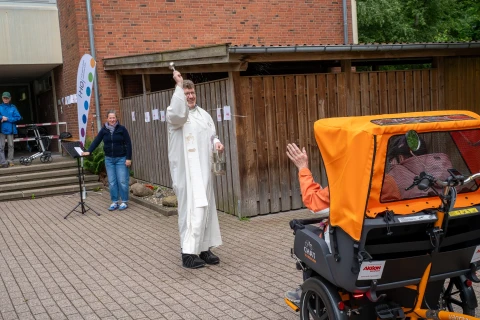 Ein Mann in cremefarbigem liturgischem Gewand steht auf einem gepflasterten Platz und hebt mit einer Hand ein Aspergill, w&auml;hrend er mit der anderen ein Metallgef&auml;&szlig; h&auml;lt. Rechts im Vordergrund ein orangener Fahrrad-Rikscha-Sitz mit einer Person darin, die einen Arm hebt. Im Hintergrund links ein Banner, eine Treppe und zwei weitere Personen. Holzwand und Backsteingeb&auml;ude im Hintergrund.