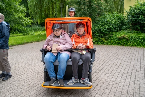Zwei Personen, beide mit Fahrradhelmen, sitzen nebeneinander in einer orangefarbenen Fahrradrikscha. Eine Person h&auml;lt eine Papiert&uuml;te. Hinter ihnen steht eine dritte Person, ebenfalls mit Fahrradhelm, die die Rikscha lenkt. Die Szene spielt auf einem gepflasterten Weg mit gr&uuml;nem Busch- und Baumbewuchs im Hintergrund. Links im Bild ist der Teil einer vierten Person erkennbar.