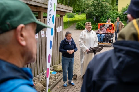 Person im wei&szlig;en liturgischen Gewand steht an einem Notenst&auml;nder und spricht. Neben ihr eine Frau in blauer Jeans und marineblauem Schal. Links im Bild eine Fahne mit Logo und bunten Punkten. Im Hintergrund zwei Menschen in einem orangefarbenen Rad mit Sitzkabine und weitere Personen vor einem Holzzaun und gr&uuml;nem Garten.