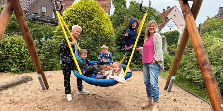Holzschaukel mit gelben Seilen und blauer Nestschaukel auf einem sandigen Spielplatz. F&uuml;nf Kinder sitzen und liegen in der Schaukel. Zwei Erwachsene stehen daneben, eine links, die andere rechts. Hintergrund mit gr&uuml;nen B&uuml;schen, B&auml;umen, Z&auml;unen und H&auml;usern mit roten D&auml;chern.