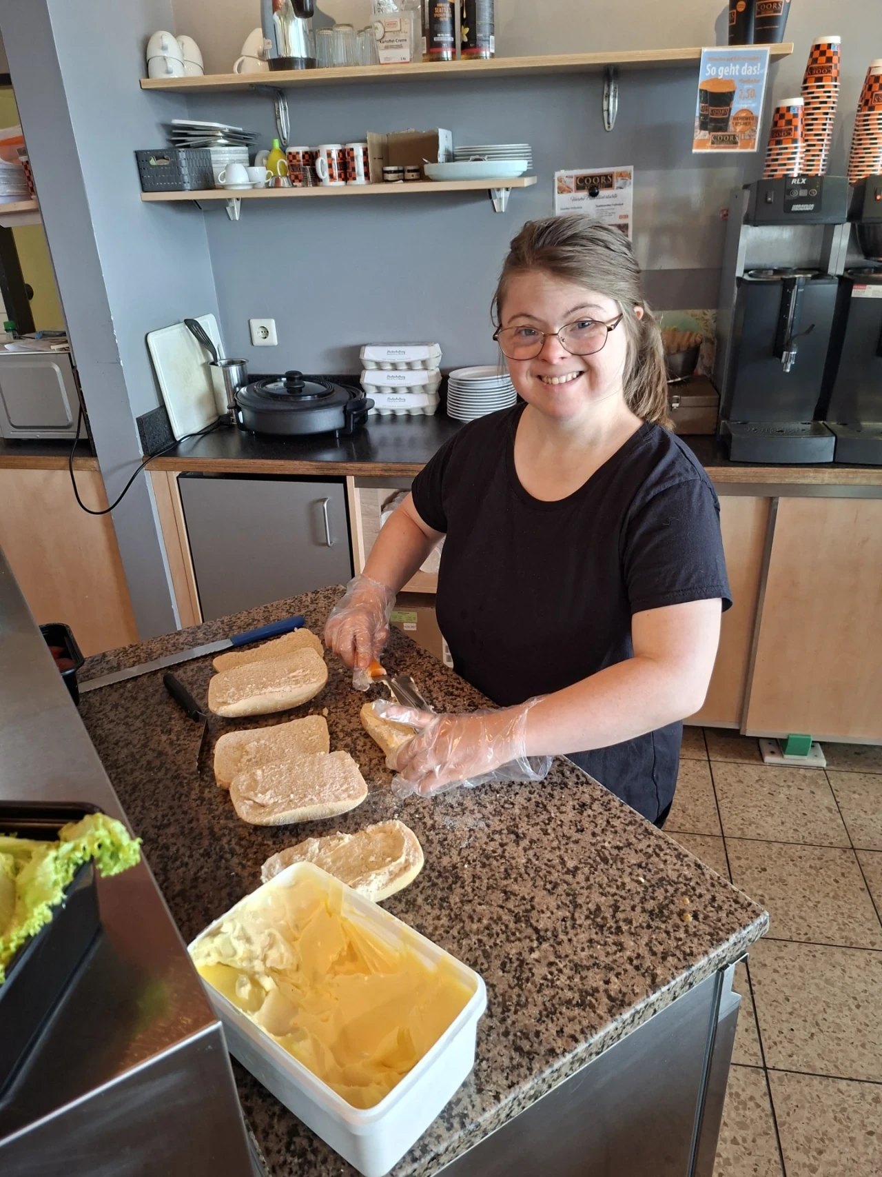 Frau mit schwarzem Oberteil, Einweghandschuhen und Brille steht an einer Arbeitsplatte in einer K&uuml;che und streicht helle Br&ouml;tchenh&auml;lften mit Butter. Links ein Beh&auml;lter mit Butter und gr&uuml;nem Salat, im Hintergrund Regale mit Tassen und K&uuml;chenutensilien.
