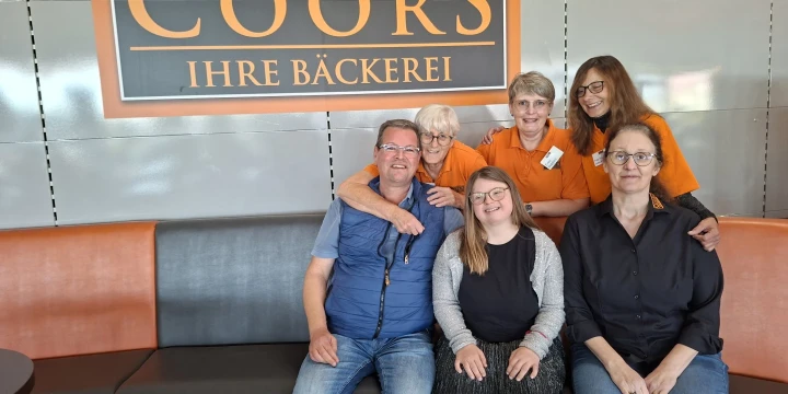 Sechs Personen sitzen auf einer schwarzen Bank vor einem Schild mit der Aufschrift &bdquo;Coors &ndash; Ihre B&auml;ckerei&ldquo;. Drei der Personen tragen orangefarbene Kleidung, die anderen drei sind in dunklen Farben gekleidet. Die Gruppe sitzt eng beisammen und l&auml;chelt in die Kamera.