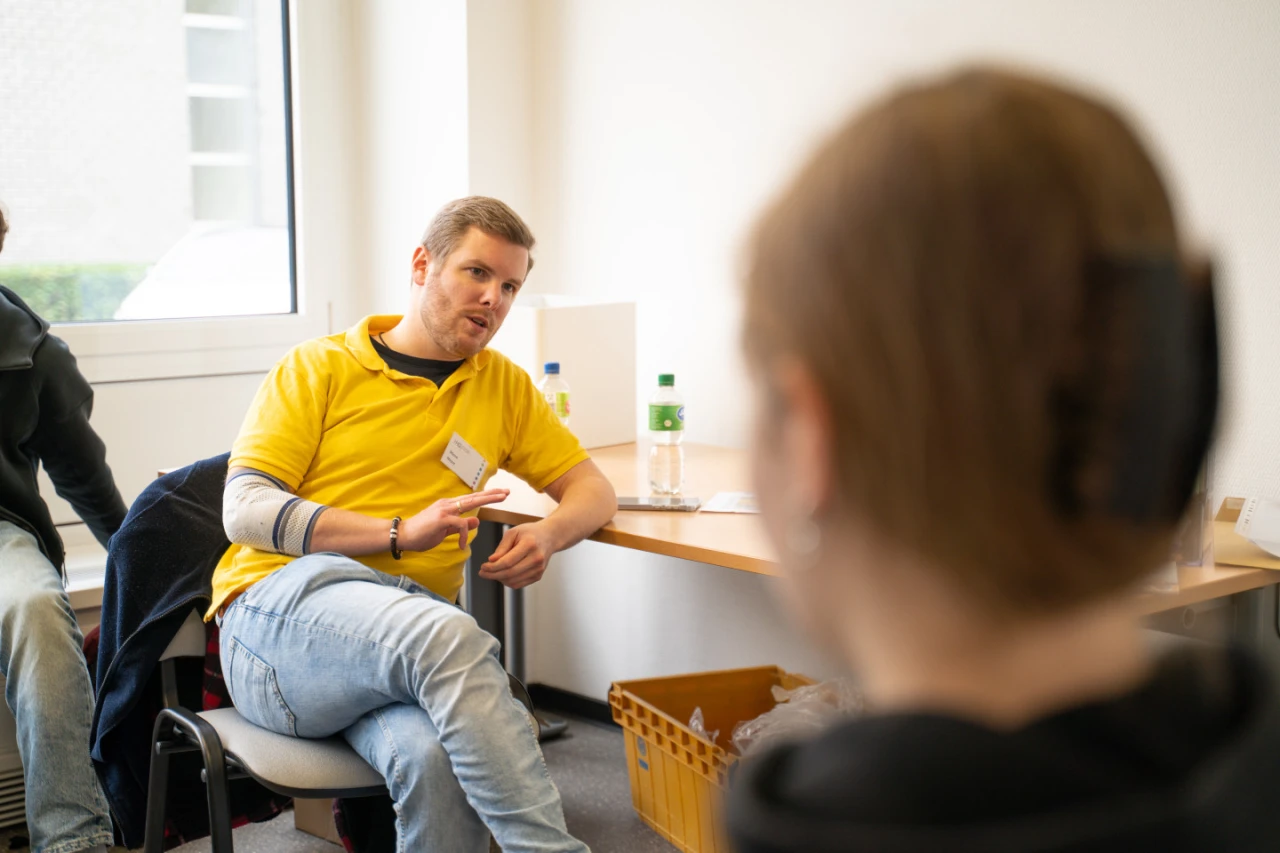 Ein Mann in gelbem Polohemd mit Namensschild sitzt auf einem Stuhl in einem hellen Raum und gestikuliert, w&auml;hrend er mit einer Person im Vordergrund, die im unscharfen Fokus ist, spricht. Rechts neben ihm ein Tisch mit einer Wasserflasche, Gl&auml;sern und einer gelben Kiste. Links ein Fenster mit Blick auf Gr&uuml;nfl&auml;chen.