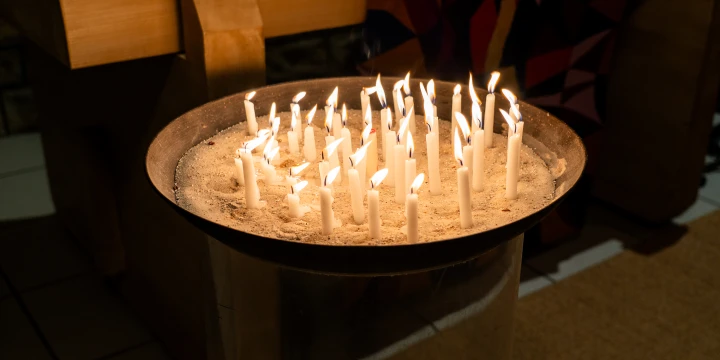 Metallene Schale, gef&uuml;llt mit wei&szlig;em Sand, in die mehrere brennende wei&szlig;e Kerzen gesteckt sind. Die Schale steht auf einem durchsichtigen Glaszylinder, im Hintergrund Holzm&ouml;bel und bunte Stoffe.