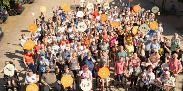 Eine gro&szlig;e Gruppe von Menschen unterschiedlicher Altersgruppen in einem Au&szlig;enbereich, stehend oder in Rollst&uuml;hlen. Die Personen halten orangefarbene und wei&szlig;e Schilder mit der Aufschrift "HO!" in die Luft. Einige tragen wei&szlig;e T-Shirts mit dem gleichen Aufdruck. Pflastersteine und geparkte Autos im Hintergrund.