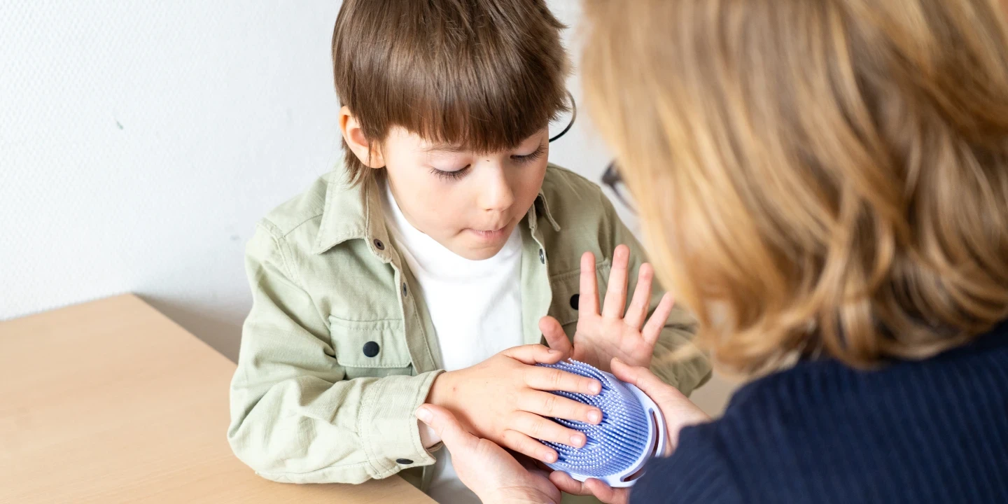 Junge mit braunen Haaren in gr&uuml;nem Hemd und wei&szlig;em T-Shirt sitzt an einem hellen Holztisch und ber&uuml;hrt mit beiden H&auml;nden einen blauen, runden Ball mit Noppen. Frau mit blondem Haar und dunkelblauer Kleidung h&auml;lt den Ball vor ihm.