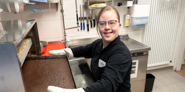 Person in dunkler Arbeitskleidung und wei&szlig;en Hitzeschutzhandschuhen nimmt ein mit braunem Kuchen gef&uuml;lltes Backblech aus einem Backofen in einer professionellen K&uuml;che. Im Hintergrund wei&szlig;e geflieste W&auml;nde, K&uuml;chenregale mit Utensilien, Aufbewahrungsbeh&auml;lter und ein Edelstahl-Arbeitsbereich.
