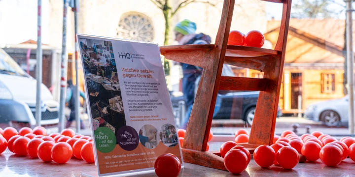 Roter Holzstuhl auf gl&auml;nzendem Boden, umgeben von zahlreichen roten Plastikb&auml;llen. Rechts neben dem Stuhl ein Infoposter auf einem Aufsteller mit dem Titel "Zeichen setzen gegen Gewalt", Text und Bildern von Menschen in einer Gemeinschaft. Im Hintergrund verschwommen Stra&szlig;enansicht mit Autos, B&auml;umen und einem Geb&auml;ude mit rundem Fenster.