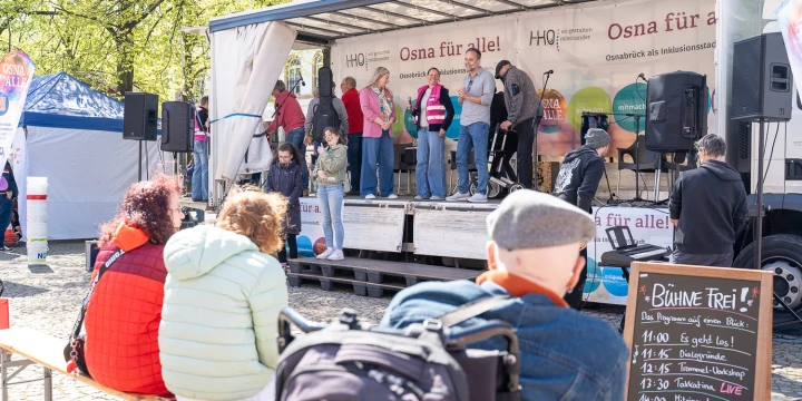 Personengruppe auf der B&uuml;hne eines offenen Transporters unter dem Banner &bdquo;Osna f&uuml;r alle!&ldquo;. Im Vordergrund drau&szlig;en sitzende Personen auf B&auml;nken, vor ihnen eine Tafel mit Programmpunkten: 11 Uhr Gespr&auml;chsrunde, 12 Uhr Dialoge live, 13 Uhr Tanzworkshop, 14 Uhr Theater live, 14:30 Akustischer Chor live, 15 Uhr Trommeln. Umgebung mit Pavillons, B&auml;umen und sonnigem Wetter.