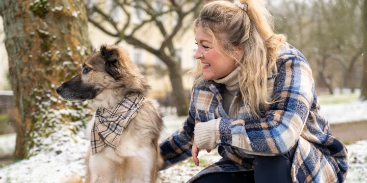 Eine Mischung aus kurzhaarigem und langhaarigem Hund mit sandfarbenem Fell und schwarzer Schnauze, tr&auml;gt ein kariertes Tuch um den Hals, sitzt im Schnee neben einer Person in blau kariertem Mantel und hellbeigem Pullover, die knieend den Hund streichelt; im Hintergrund verschneite B&auml;ume und ein Geb&auml;ude.
