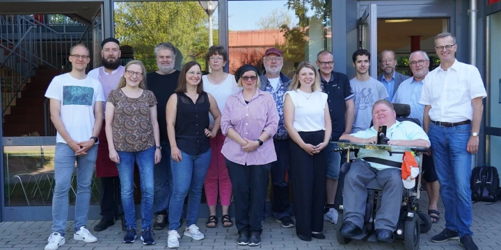 Eine Gruppe von elf stehenden Menschen und einer Person in einem elektrischen Rollstuhl vor einem Geb&auml;ude mit Glasfassade und roten Elementen. Im Hintergrund ein Treppenaufgang, ein Baum und leicht sichtbarer Zaun. Der Boden besteht aus grauen Pflastersteinen; im Vordergrund ist eine Rasenfl&auml;che.