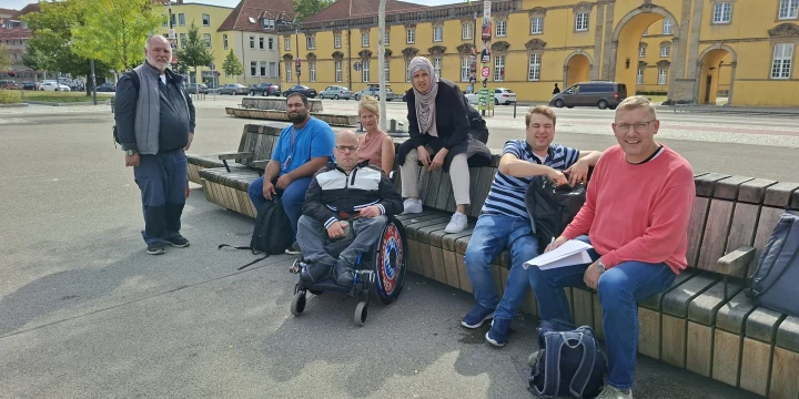 Sechs Personen sitzen und eine Person steht auf einem &ouml;ffentlichen Platz auf Holzsitzb&auml;nken im Halbkreis. Eine Person im Rollstuhl sitzt mittig. Im Hintergrund gelbliche Geb&auml;ude, B&auml;ume, geparkte Autos und Stra&szlig;enlaternen.