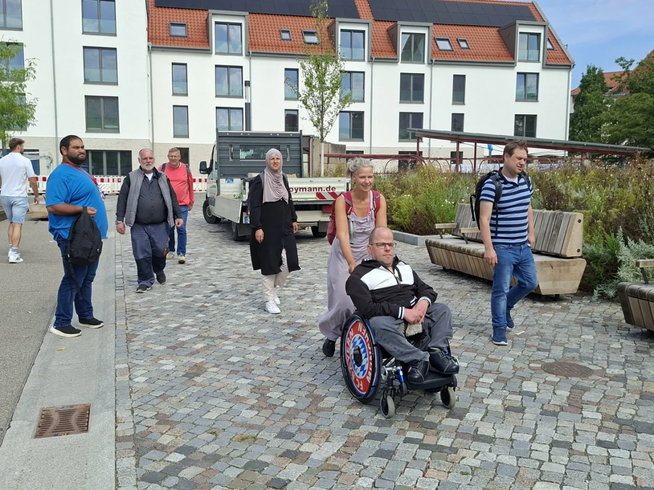 Gruppe von sieben Menschen auf einem gepflasterten Gehweg zwischen modernen wei&szlig;en Geb&auml;uden mit roten Ziegeld&auml;chern. Eine Person im Rollstuhl mit einem markanten Radmuster wird geschoben. Neben und hinter ihnen gehen weitere Personen. Auf der rechten Seite Pflanzenbeete und Holzb&auml;nke, links ein geparktes Fahrzeug.