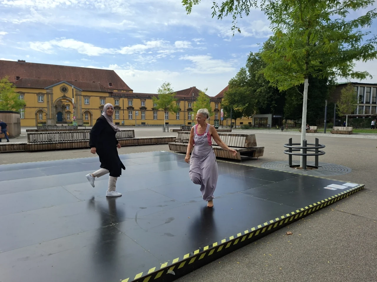 Zwei Personen tanzen auf einer schwarzen Outdoor-B&uuml;hne mit gelb-schwarz markierten Kanten. Eine Person tr&auml;gt ein schwarzes, langes Kleid mit wei&szlig;em Kopftuch und Sneakern, die andere einen grauen Jumpsuit und tanzt barfu&szlig;. Im Hintergrund ein gro&szlig;er Platz mit B&auml;umen, Holzb&auml;nken und gelben Geb&auml;uden mit roten D&auml;chern.