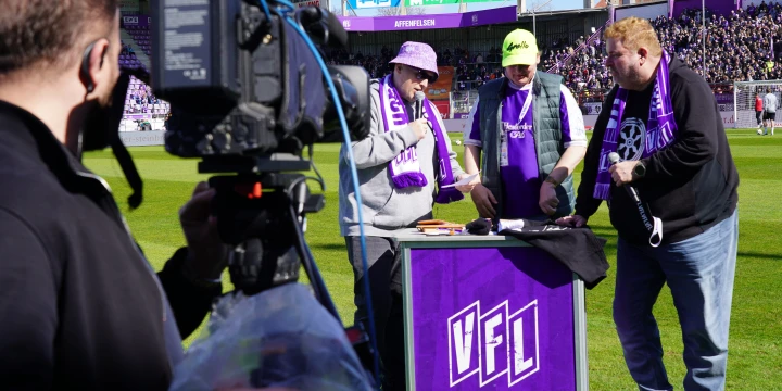 Drei Personen mit lila VfL-Schals stehen an einem Tisch mit lila VfL-Logo, umgeben von Equipment. Links ein Kameramann mit gro&szlig;er Videokamera. Im Hintergrund ein Fu&szlig;ballstadion, Trib&uuml;nen mit Zuschauern und Werbebanner. Sonnige Wetterbedingungen.
