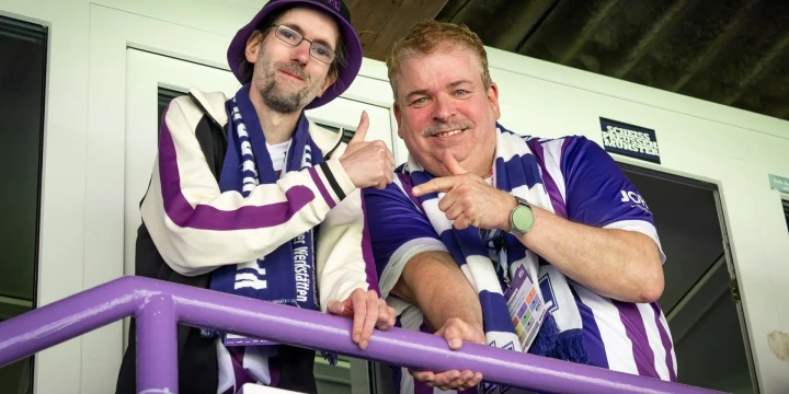 Zwei Personen stehen auf einem Balkon mit einem violetten Gel&auml;nder. Beide tragen Kleidung in wei&szlig;en und violetten Farben sowie Fan-Schals. Die linke Person tr&auml;gt einen Hut und eine Jacke, die rechte ein gestreiftes Trikot und eine Armbanduhr. Beide Personen gestikulieren und wirken fr&ouml;hlich. Im Hintergrund befinden sich eine wei&szlig;e T&uuml;r und eine Decke mit sichtbaren Metallstreben.