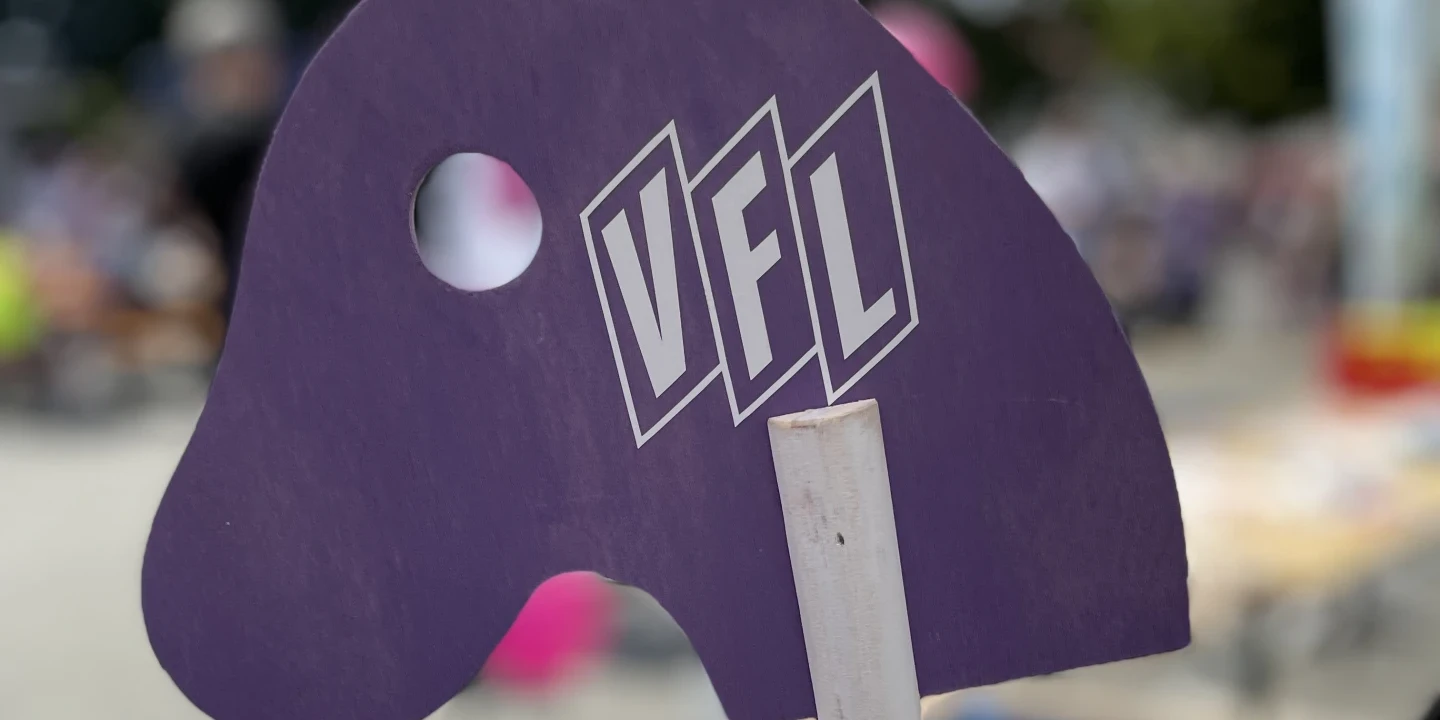 Lila Holzfigur in Form eines Pferdekopfs mit einem ausgestanzten Kreis f&uuml;r das Auge und der Aufschrift "VFL" in wei&szlig;en, gro&szlig;en Buchstaben. Befestigt auf einem Holzst&auml;nder, dieser steht auf einem hellen runden Tisch. Rechts ein Laptop, links unscharf ein Blatt Papier. Hintergrund unscharf mit Personen und B&auml;umen.