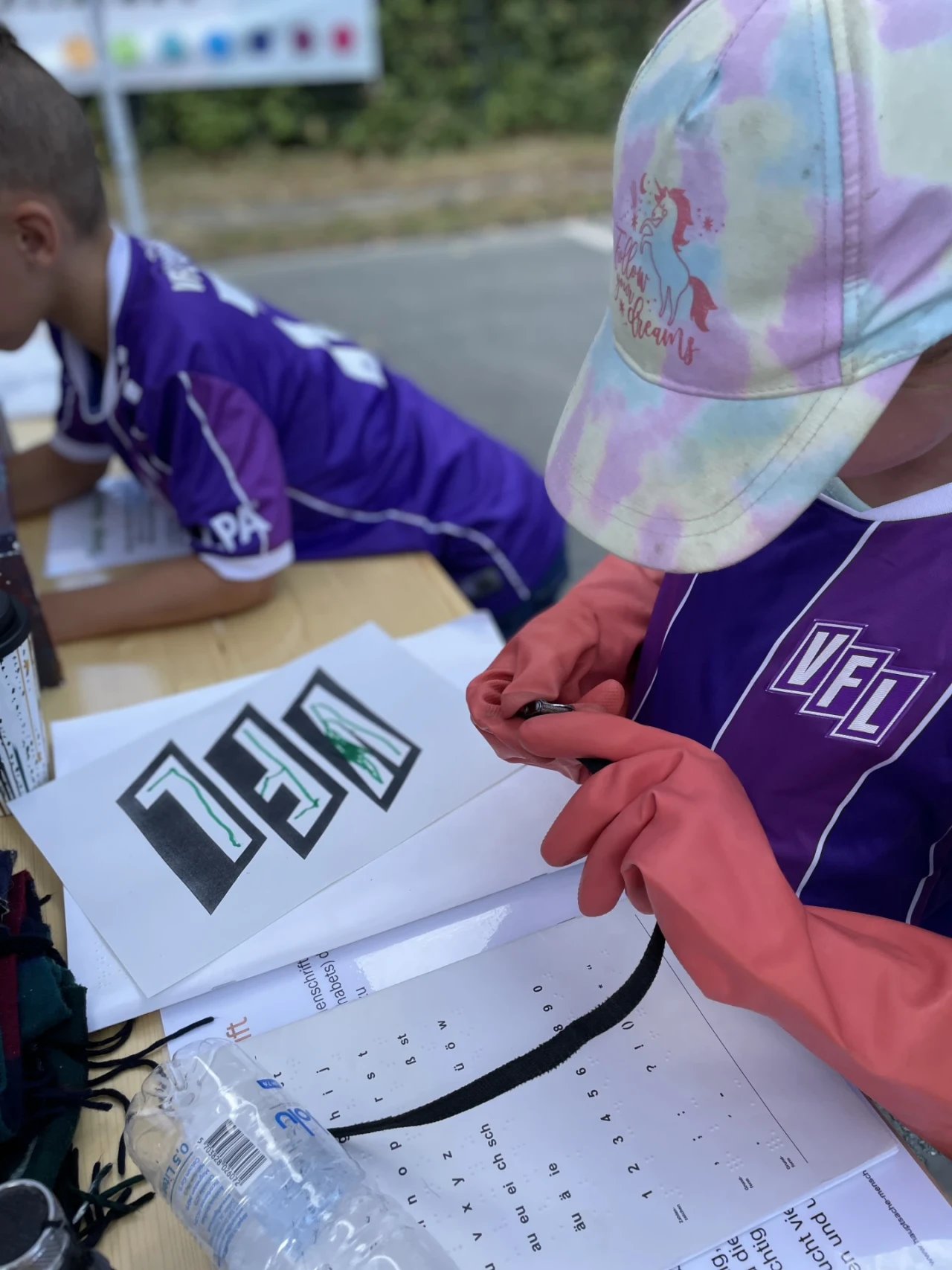 Kind in lila Trikot mit wei&szlig;em &bdquo;VFL&ldquo;-Logo, pinken Gummihandschuhen und bunter Kappe bemalt Papier mit schwarzem Schriftzug &bdquo;VFL&ldquo;, umgeben von Bastelmaterialien, Plastikflasche und Arbeitsunterlagen auf einem Holztisch. Im Hintergrund weiteres Kind in lila Trikot und unscharfer Au&szlig;enbereich.