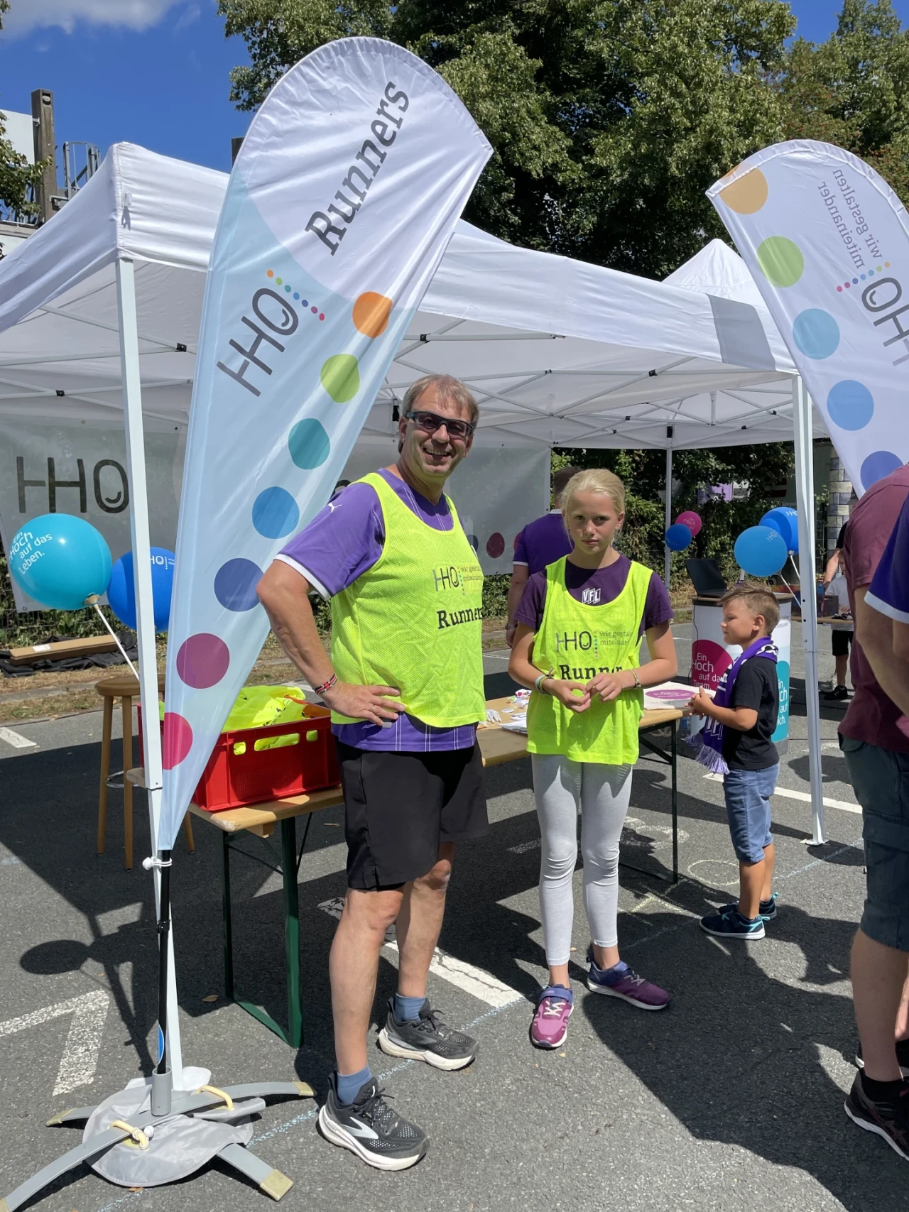 Zwei Personen in gelben Warnwesten mit lila T-Shirts darunter, beschriftet mit "HHO Runners", stehen unter einem wei&szlig;en Pavillon. Links und rechts von ihnen stehen wei&szlig;e Beachflags mit bunten Kreisen und der Aufschrift "HHO Runners". Im Hintergrund sind Luftballons, ein Kind, ein Tisch mit Materialien und ein weiterer Pavillon zu sehen.