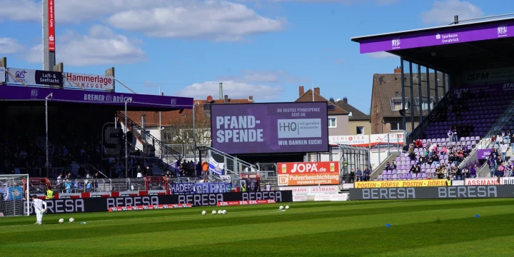 Gr&uuml;ner Fu&szlig;ballrasen mit mehreren verstreuten Fu&szlig;b&auml;llen, umgeben von Trib&uuml;nen. Trib&uuml;ne links in Schwarz und Violett mit Zuschauerbereich. Rechte Trib&uuml;ne teilweise &uuml;berdacht, ebenfalls in Violett. Auf Werbetafel steht &bdquo;Pfandspende &ndash; Die n&auml;chste Pfandspende kommt dem Fanshop zugute &ndash; Wir danken euch!&ldquo;. Klarer Himmel mit vereinzelten wei&szlig;en Wolken.