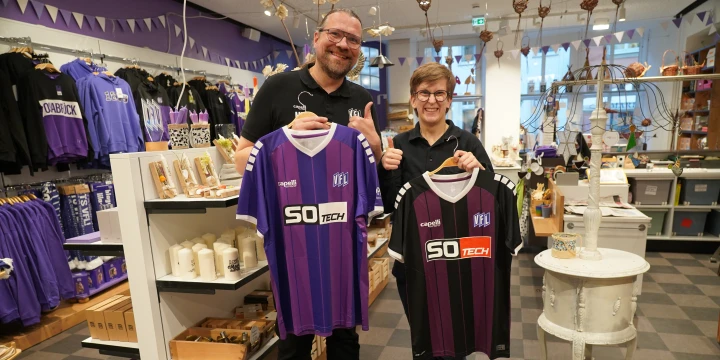 Eine gr&ouml;&szlig;ere und eine kleinere Person halten je ein Fu&szlig;balltrikot hoch. Links ein lila Trikot, rechts ein schwarz-lila gestreiftes Trikot, beide mit der Aufschrift "SO TECH". Hintergrund: Gesch&auml;ft mit Regalen voller Fanartikel in Lila und Wei&szlig;, sowie Dekoartikel auf einem rautenf&ouml;rmigen Boden.