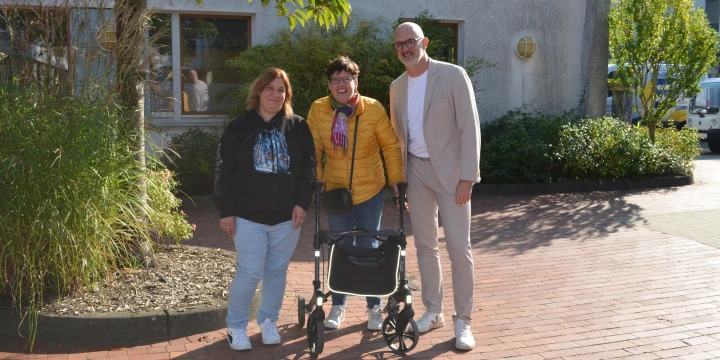 Drei Personen stehen auf einem ziegelroten Pflasterweg vor einem Geb&auml;ude mit grauer Fassade, Fenstern und umgebender Gr&uuml;nbepflanzung. Eine Person tr&auml;gt eine gelbe Jacke und nutzt einen schwarzen Rollator, daneben steht eine Person in sportlich-eleganter Kleidung und eine in dunkler Jacke mit hellblauer Jeans.