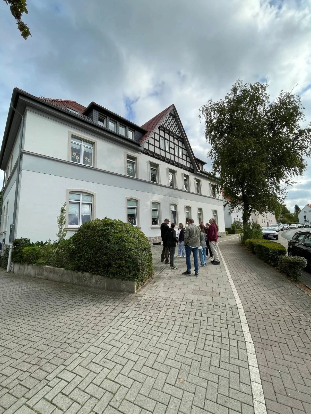 Breites, zweist&ouml;ckiges Geb&auml;ude mit wei&szlig;er Fassade und einem Fachwerkgiebel in der Mitte, kombiniert mit einem roten Ziegeldach. Mehrere Fensterreihen, einige mit Dekorationen. Davor stehen sieben Personen in einer Gruppe, darunter eine Frau in rotem Mantel. Rechts Baum und Gehweg aus Pflastersteinen mit parkenden Autos am Stra&szlig;enrand.