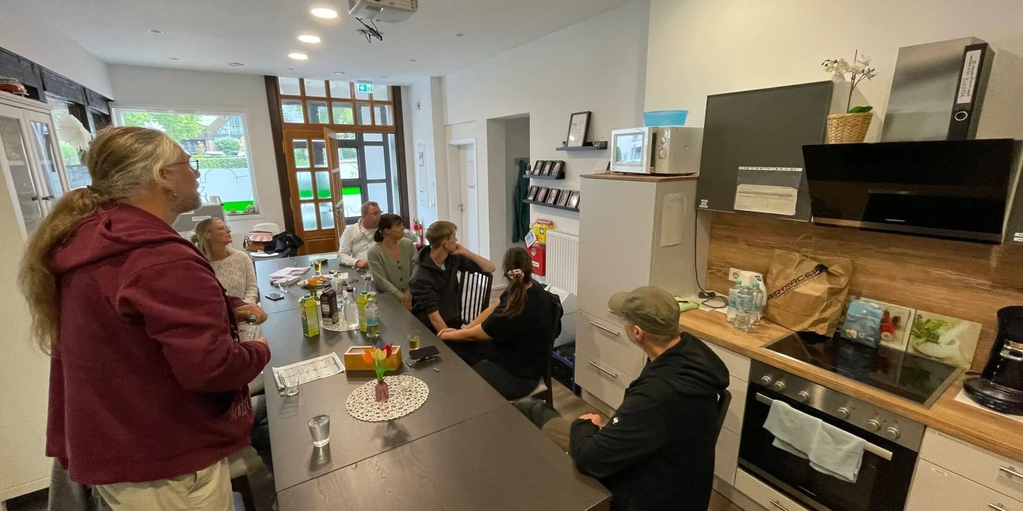 Personengruppe in moderner K&uuml;che mit Holzboden und wei&szlig;en W&auml;nden; langer Esstisch mit Getr&auml;nken und einer Vase mit Blumen in der Mitte; links stehen zwei Personen, rechts sitzen f&uuml;nf Personen auf St&uuml;hlen; im rechten Bildbereich Einbauk&uuml;che mit Backofen, Herd, Mikrowelle und Pflanzen auf oberem Regal.