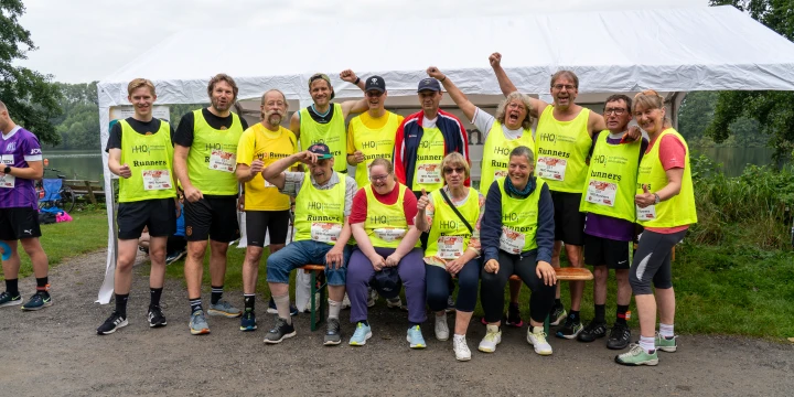 Eine Gruppe von 13 Personen in gelben Laufwesten mit der Aufschrift &bdquo;HHD Runners&ldquo;, einige tragen zus&auml;tzliche T-Shirts oder Sportkleidung. Sie stehen und sitzen vor einem wei&szlig;en Pavillon, umgeben von Natur, mit Gras, B&auml;umen und einem Gew&auml;sser im Hintergrund.