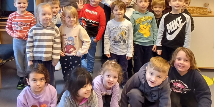 Gruppe von 16 Kindern in bunter Freizeitkleidung, teils stehend, teils kniend, in einem Klassenraum. Im Hintergrund drei Erwachsene, ein wei&szlig;es B&uuml;cherregal mit Spielmaterialien, eine analoge Wanduhr und ein Regal mit einem Globus auf der rechten Seite.