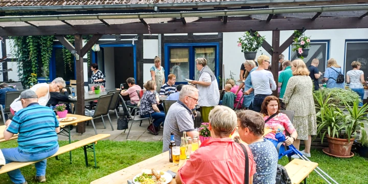 Menschen sitzen und stehen in einem Garten mit Wiese auf und um mehrere Holzb&auml;nke und -tische. Im Hintergrund ein Geb&auml;ude mit wei&szlig;en W&auml;nden, blauen Fensterrahmen, h&ouml;lzernem Vordach und Blumenampeln. Tische haben Getr&auml;nke und Speisen.