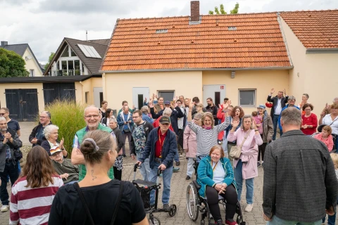 Gruppe von etwa 30 Personen auf einem gepflasterten Hof vor einem hellgelben Geb&auml;ude mit orangerotem Ziegeldach. Menschen verschiedenen Alters, teils stehend, teils sitzend, darunter eine Frau im Rollstuhl. Einige heben die H&auml;nde. Im Hintergrund weitere Geb&auml;ude mit Fenstern und G&auml;rten. Wolkiger Himmel.