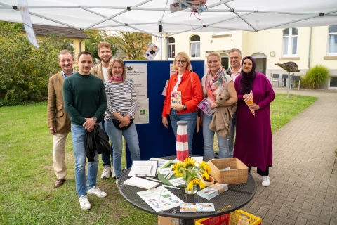 Gruppe von acht Personen unterschiedlicher Altersgruppen steht unter einem wei&szlig;en Pavillon-&Uuml;berdachung im Freien. Hinter den Personen eine dunkelblaue Pinnwand mit mehreren Papieren und Texten. Vorne ein runder Tisch mit Sonnenblumen, Flyern und anderen Dokumenten sowie einem Korb auf dem Boden. Geb&auml;ude und gr&uuml;ne Wiese im Hintergrund.