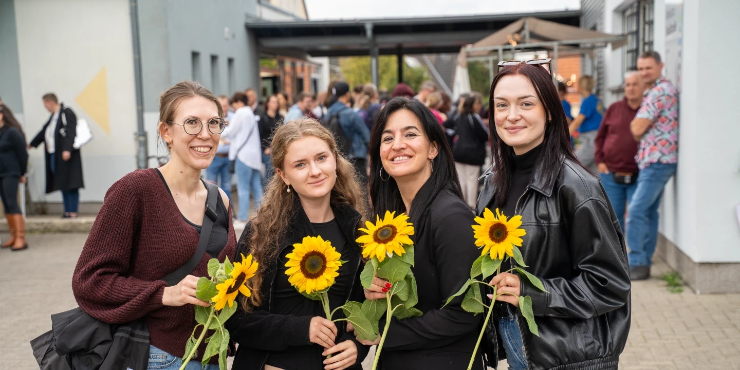 Vier l&auml;chelnde Frauen stehen nebeneinander auf einem gepflasterten Platz und halten jeweils eine Sonnenblume in der Hand. Zwei tragen Brillen, drei tragen dunkle Oberteile; eine tr&auml;gt eine schwarze Lederjacke. Im Hintergrund eine wei&szlig;e Geb&auml;udewand, Menschen in Gruppen sowie ein &uuml;berdachter Bereich.