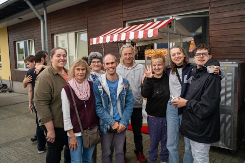 Gruppe von acht Personen unterschiedlichen Alters, die drau&szlig;en vor einem Verkaufsstand mit rot-wei&szlig; gestreiftem Dach posieren. Im Hintergrund eine Holzfassade mit Fenstern. Einige Personen l&auml;cheln, eine hebt die Hand zum Winken. Links und rechts am Rand weitere Personen teilweise sichtbar.