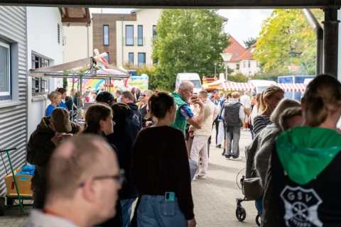 Menschenansammlung auf einem Au&szlig;enmarkt, ges&auml;umt von St&auml;nden mit gestreiften Markisen. Links ein Pavillon vor einer grauen Geb&auml;udewand. Bunte H&uuml;pfburgen und B&auml;ume im Hintergrund. Person in t&uuml;rkis-blauem Pulli mittig h&auml;lt ein Kind, umgeben von weiteren Personen.