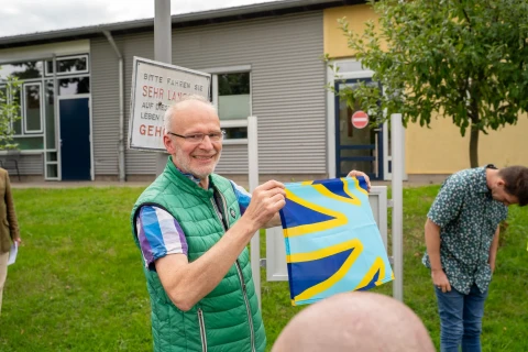 Ein Mann mit gr&uuml;ner Steppweste h&auml;lt ein kleines Tuch mit blau-gelbem Kreuzmuster in die Kamera. Im Hintergrund eine Person mit gr&uuml;n gemusterter Kleidung, ein Geb&auml;ude mit grauer Fassade, ein Baum und ein Schild mit der Aufschrift &bdquo;Bitte fahren Sie sehr langsam... Gehen&ldquo;. Im Vordergrund ein angedeuteter kahler Kopf.