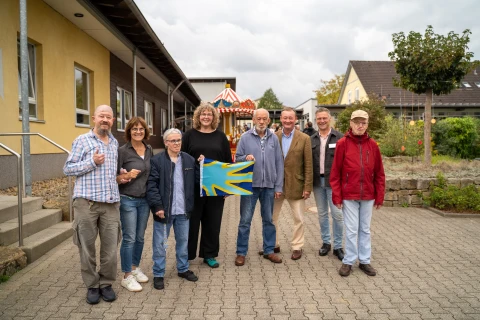 Eine Gruppe von acht Personen steht auf einem gepflasterten Hof vor einem Geb&auml;ude mit gelber und brauner Fassade. Eine Frau in der Mitte h&auml;lt ein rechteckiges, buntes Bild mit blauen und gelben Mustern. Im Hintergrund befinden sich ein kleiner Pavillon sowie ein weiteres Geb&auml;ude mit grauem Dach und umliegender Vegetation. Der Himmel ist leicht bew&ouml;lkt.